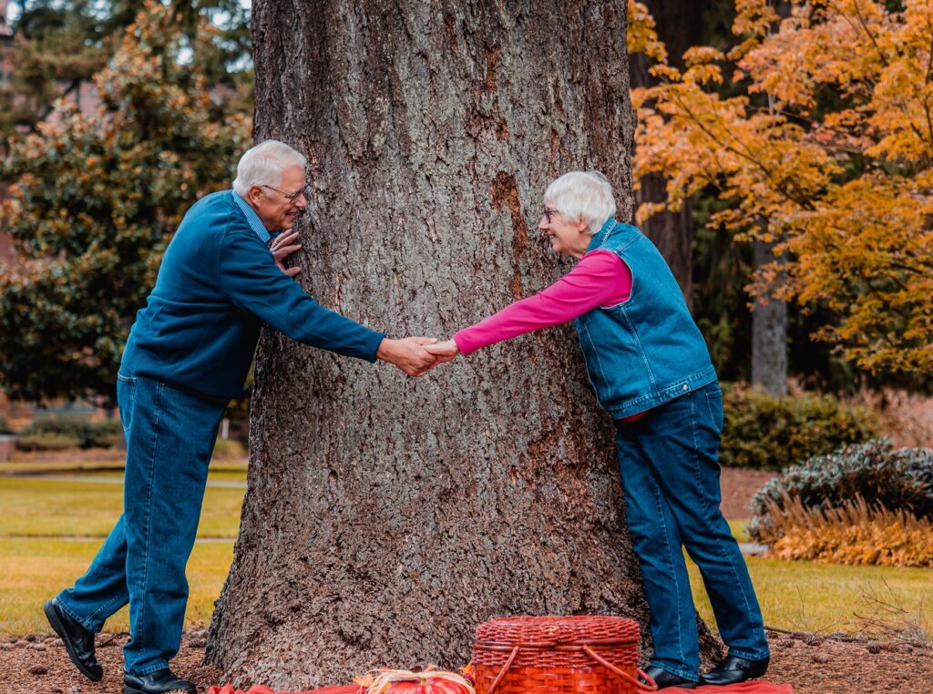 Spending Time With Your Senior Loved One This Autumn - smiling Couple holding hands in front of large tree golden leaves surround picnic